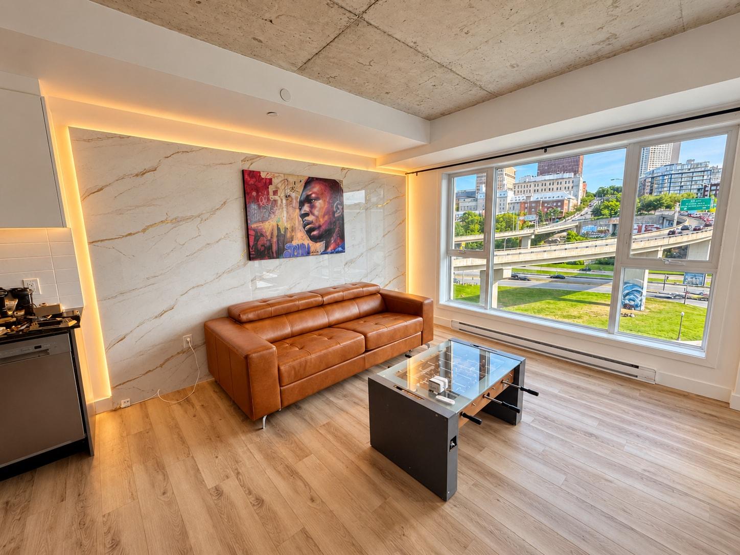Le salon : canapé de cuir cognac devant un mur de marbre rétroéclairé, table basse baby-foot et grandes fenêtres